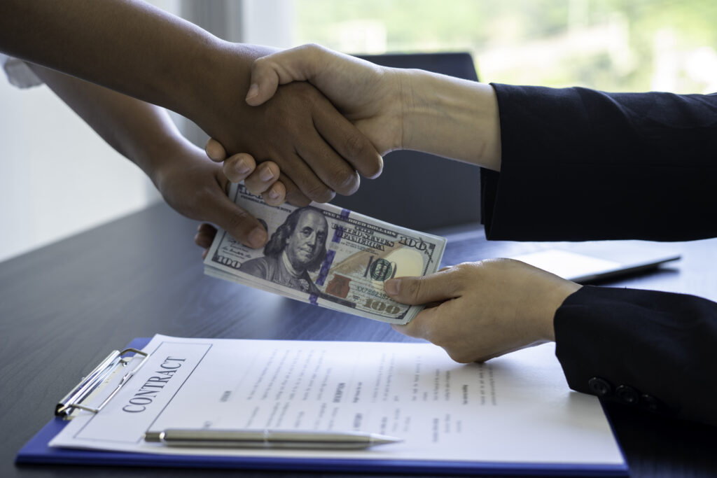 Two people shaking hands while exchanging cash over a signed contract on a desk.