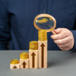 A businessman analyzing financial growth with a magnifying glass over stacked coins on ascending wooden blocks, each marked with upward arrows, symbolizing increasing income or investment returns