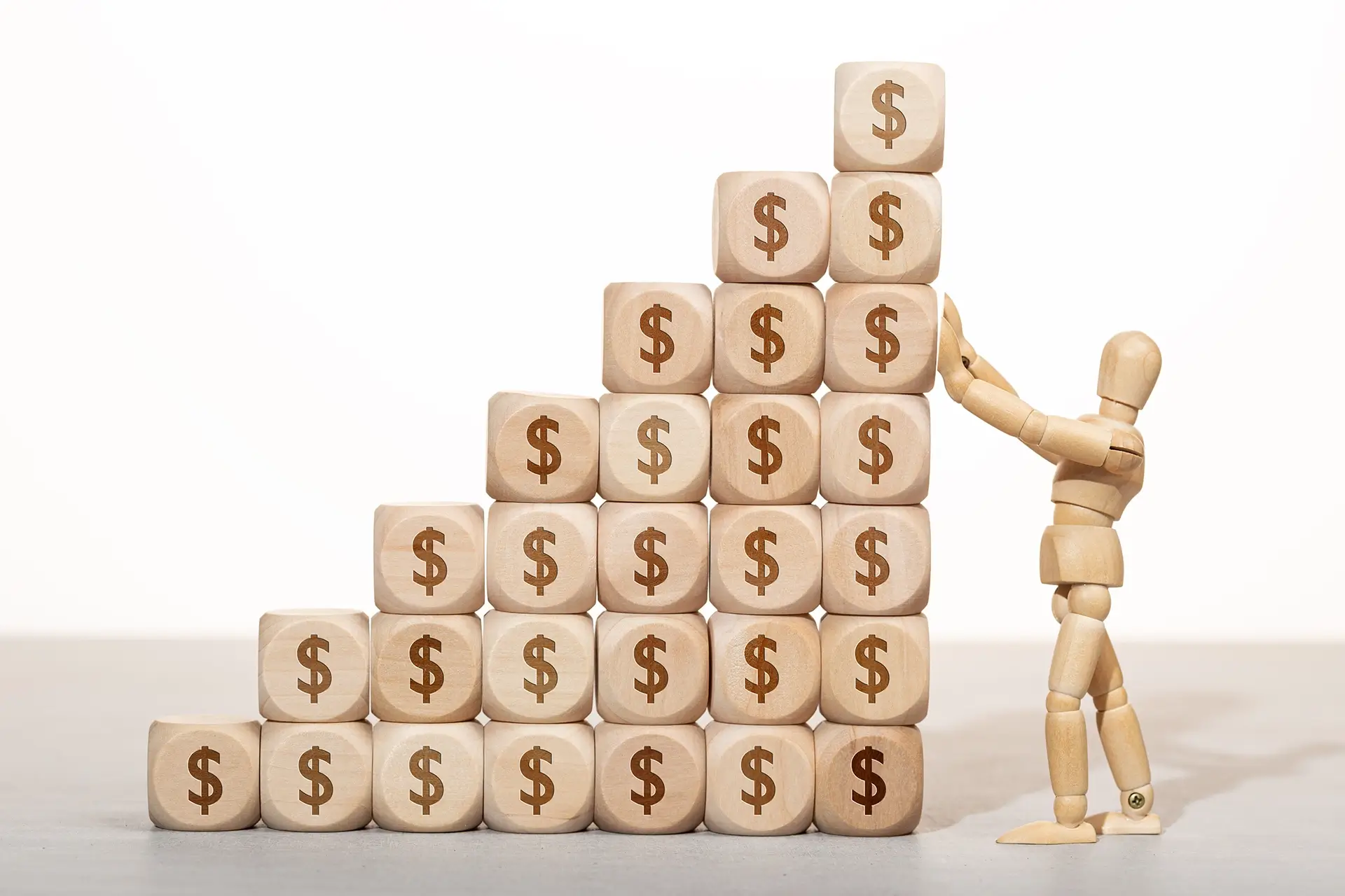 A wooden figure stands atop a stack of wooden blocks, each marked with dollar signs, symbolizing financial success.